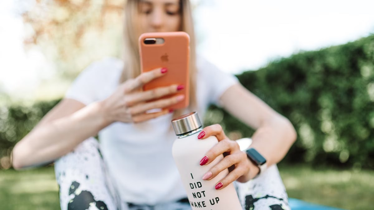 Woman outdoors using smartphone while holding a water bottle with text. Casual and relaxed setting.